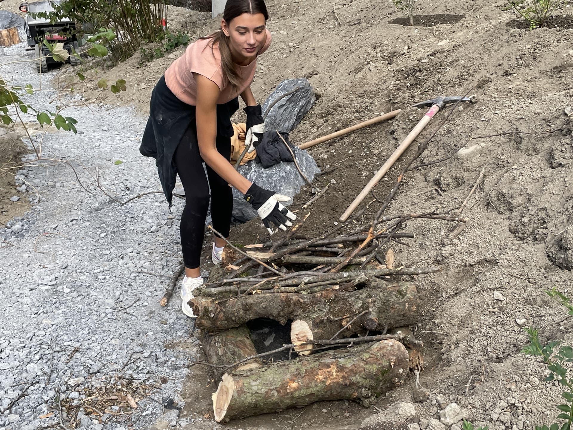 Eine junge Frau schichtet mit Gartenhandschuhen Äste aufeinander.