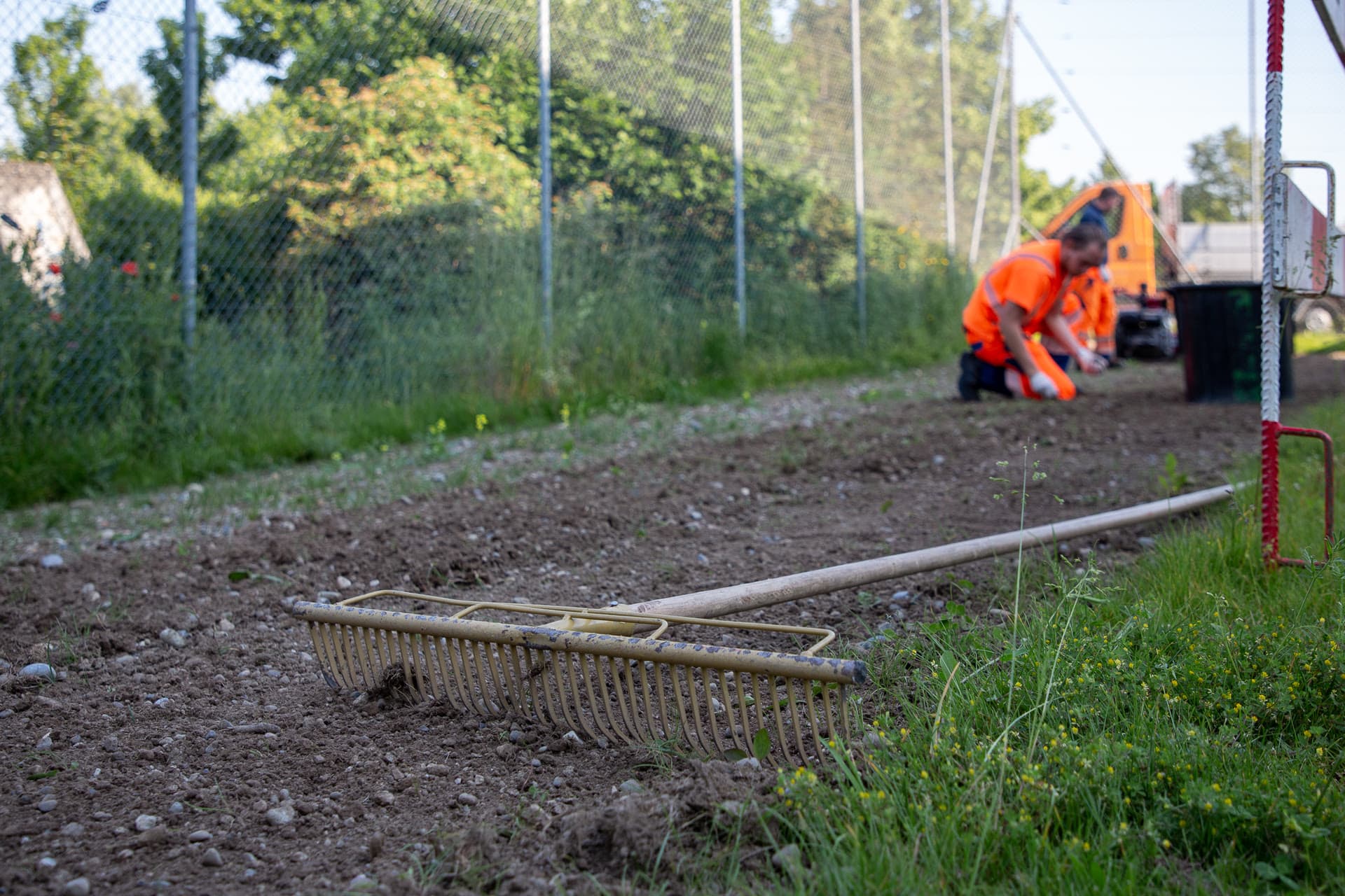 Rechen liegt auf einer Fläche, die gefräst und für eine Blumenwiese vorbereitet wurde, im Hintergrund kniet ein Werkhofmitarbeiter