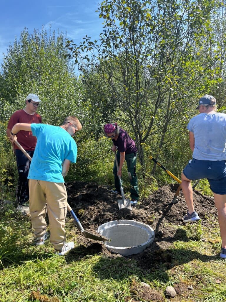 Vier Männer im Grünen versenken ein Betonrohr im Boden um eine Unkenwanne zu gestalten.