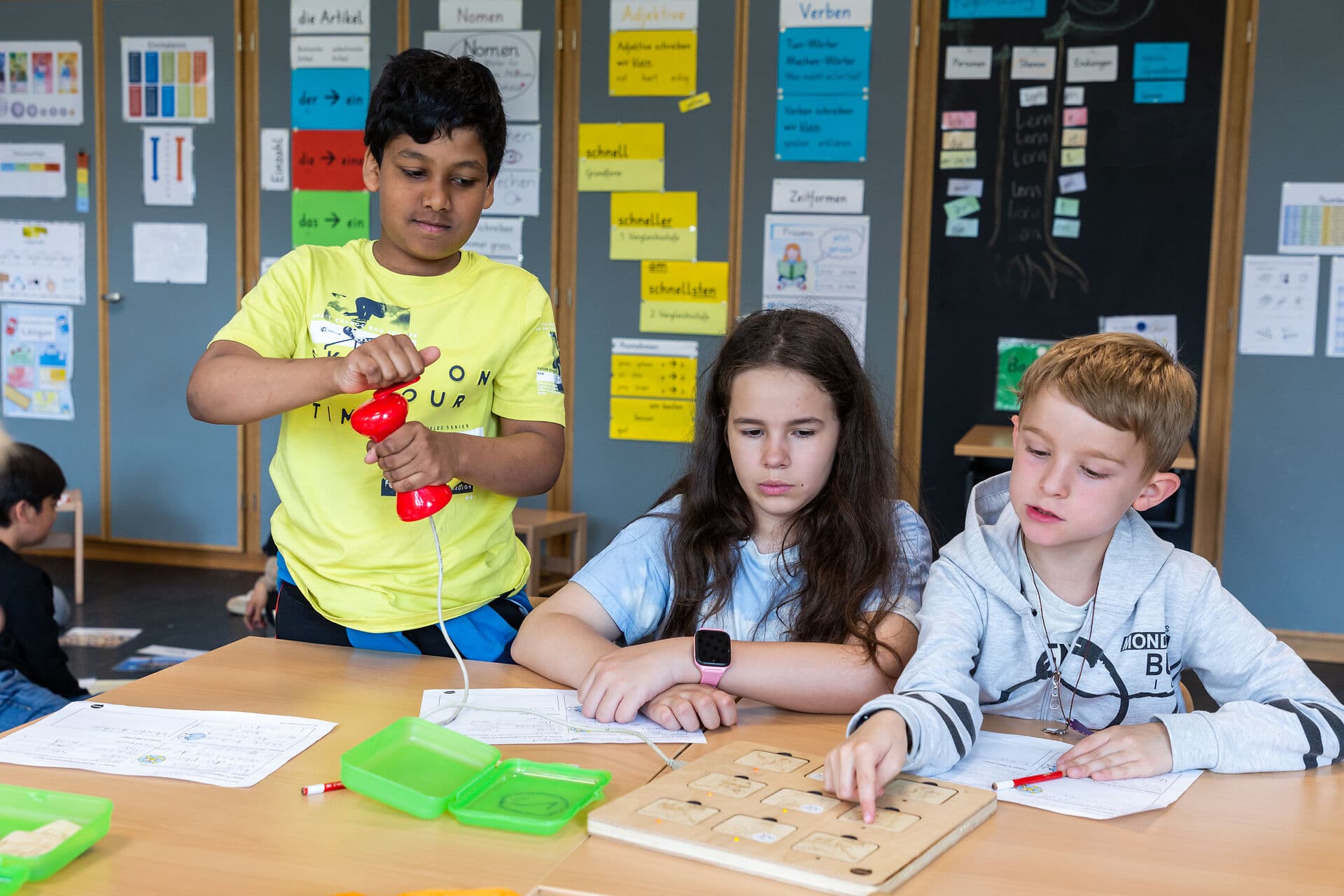 Drei Kinder sitzen an einem Tisch mit Holzspiel, auf dem mit einer roten Kurbel Lämpchen angezündet werden