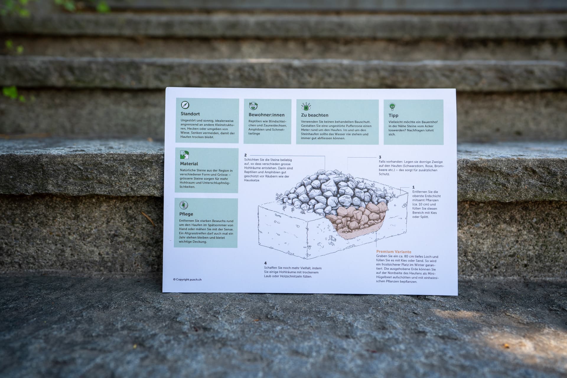 Ein Merkblatt mit einer schwarz-weiss Skizze eines Steinhaufens und Erklärungen dazu, steht an eine steinerne Treppenstufe angelehnt.